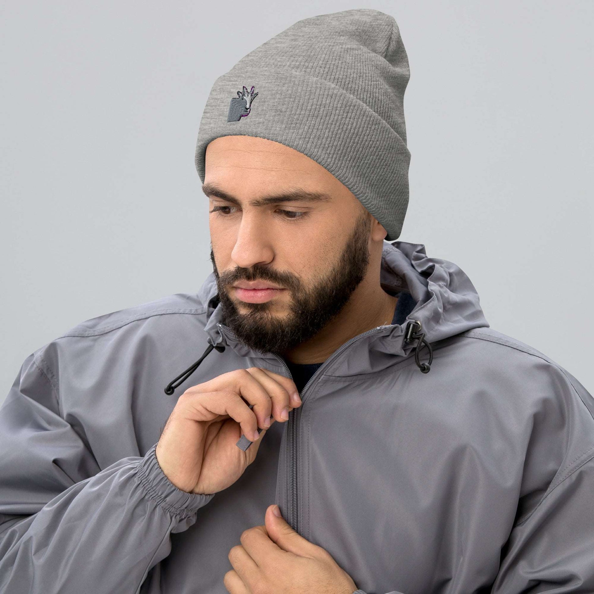 A male model wearing a high-quality light grey unisex cuffed beanie that has an embroidery of a funny mountain goat design and a running jacket.