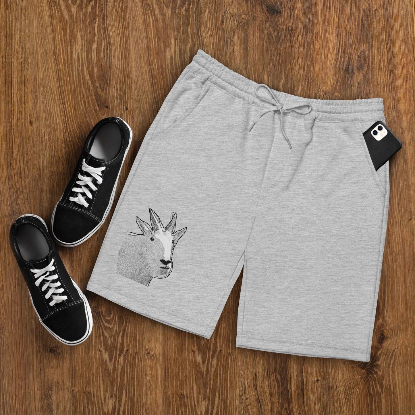 A pair of grey Men's fleece shorts designed by North American Mountain Goat Appreciation Club that features a unique funny mountain goat design, placed on a natural wooden floor beside a pair of black sneakers.