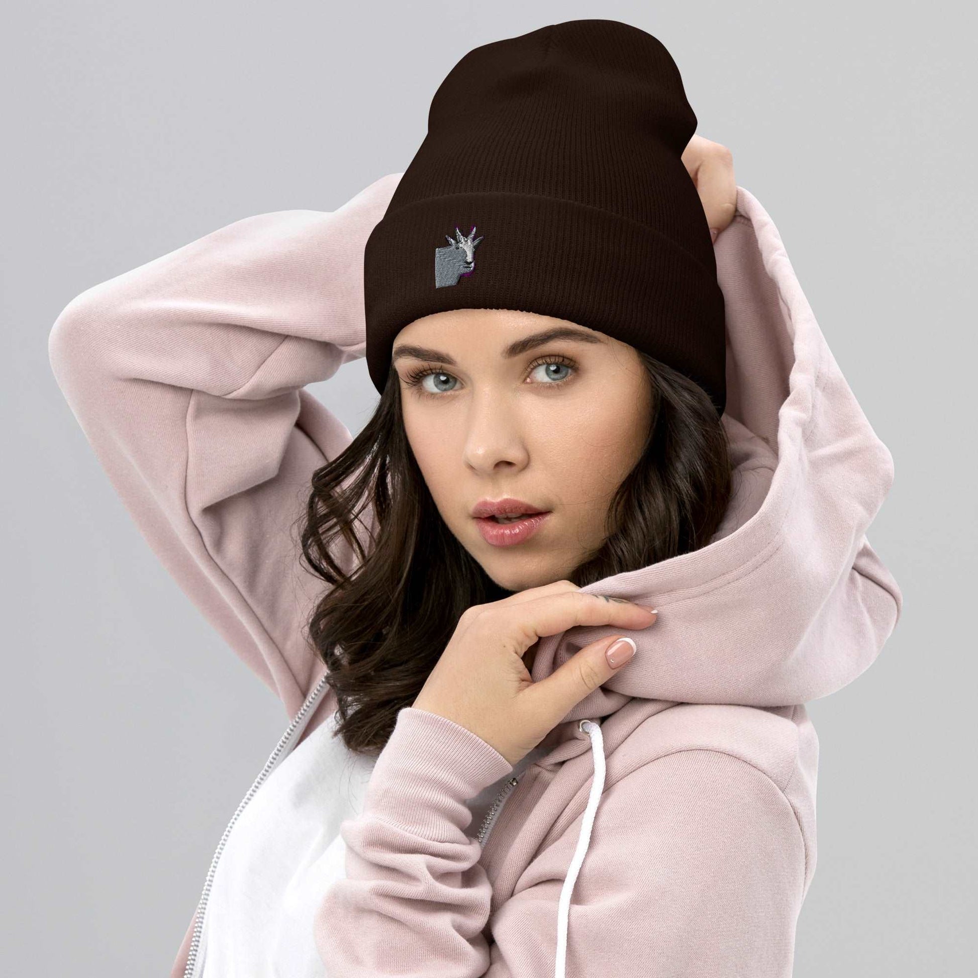 A female model wearing a high-quality brown unisex cuffed beanie that has an embroidery of a funny mountain goat design.