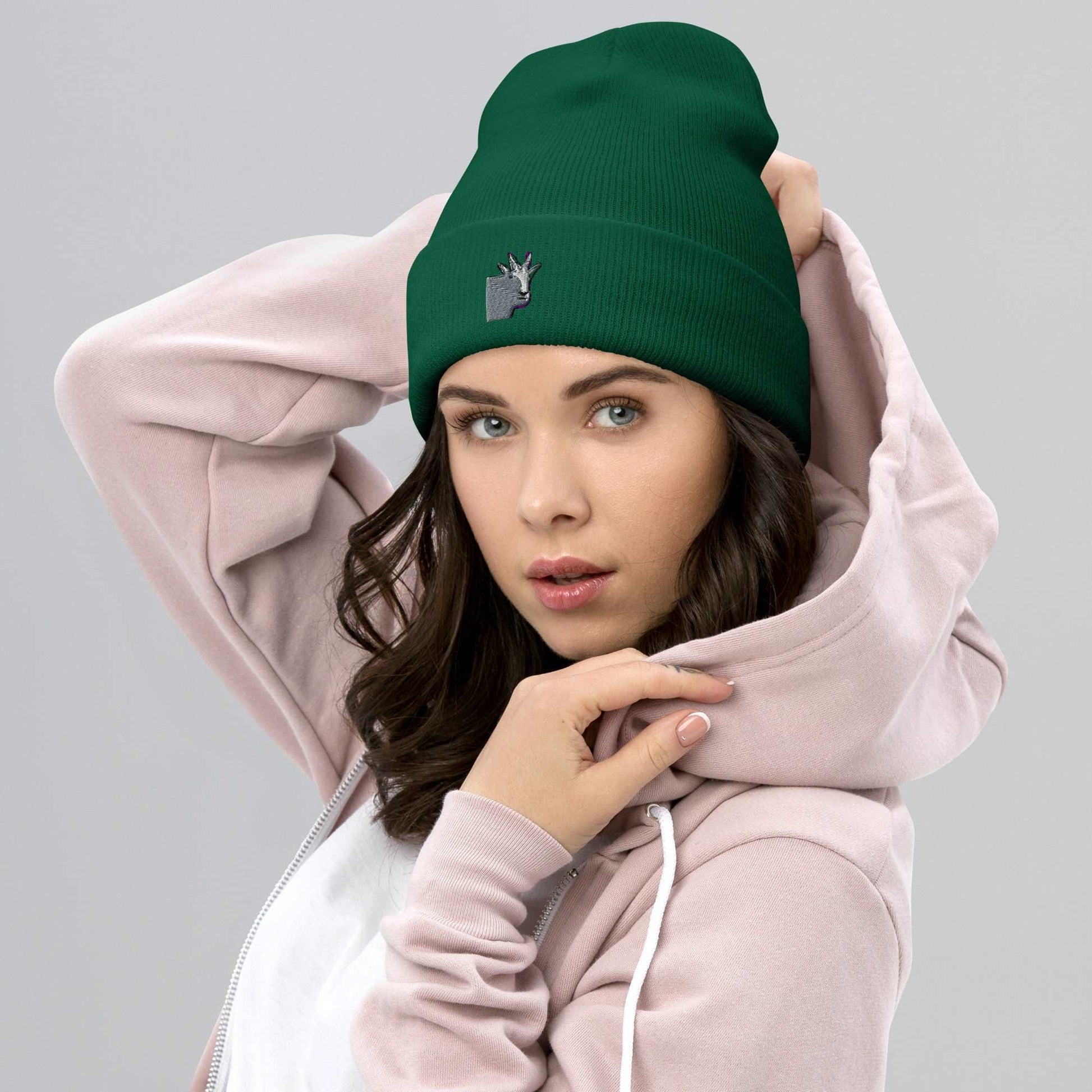 A female model wearing a high-quality spruce green unisex cuffed beanie that has an embroidery of a funny mountain goat design.