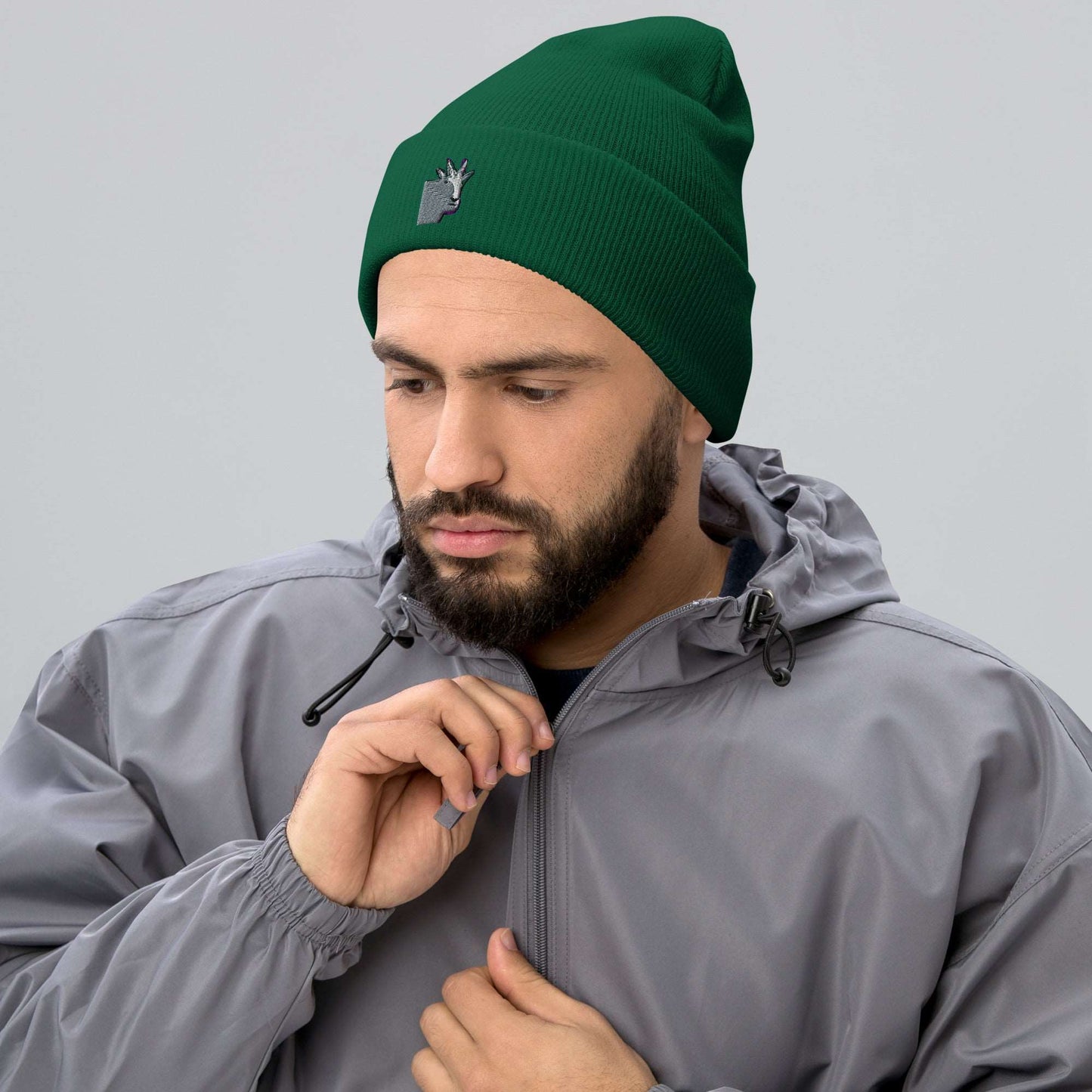 A male model wearing a high-quality spruce green unisex cuffed beanie that has an embroidery of a funny mountain goat design and a running jacket.