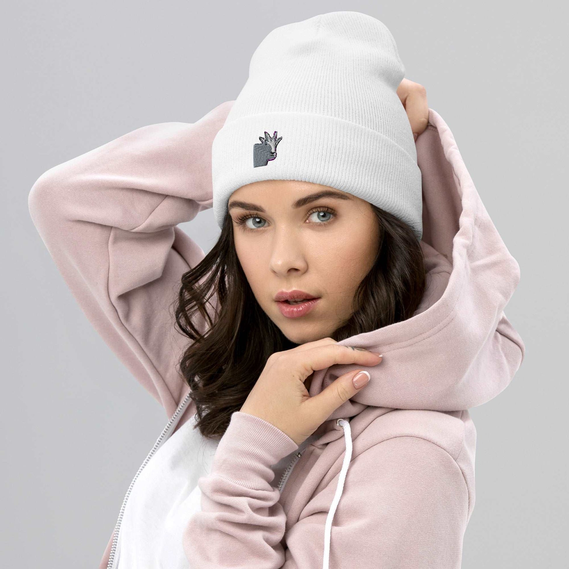 A female model wearing a high-quality white unisex cuffed beanie that has an embroidery of a funny mountain goat design and a pink hoodie.