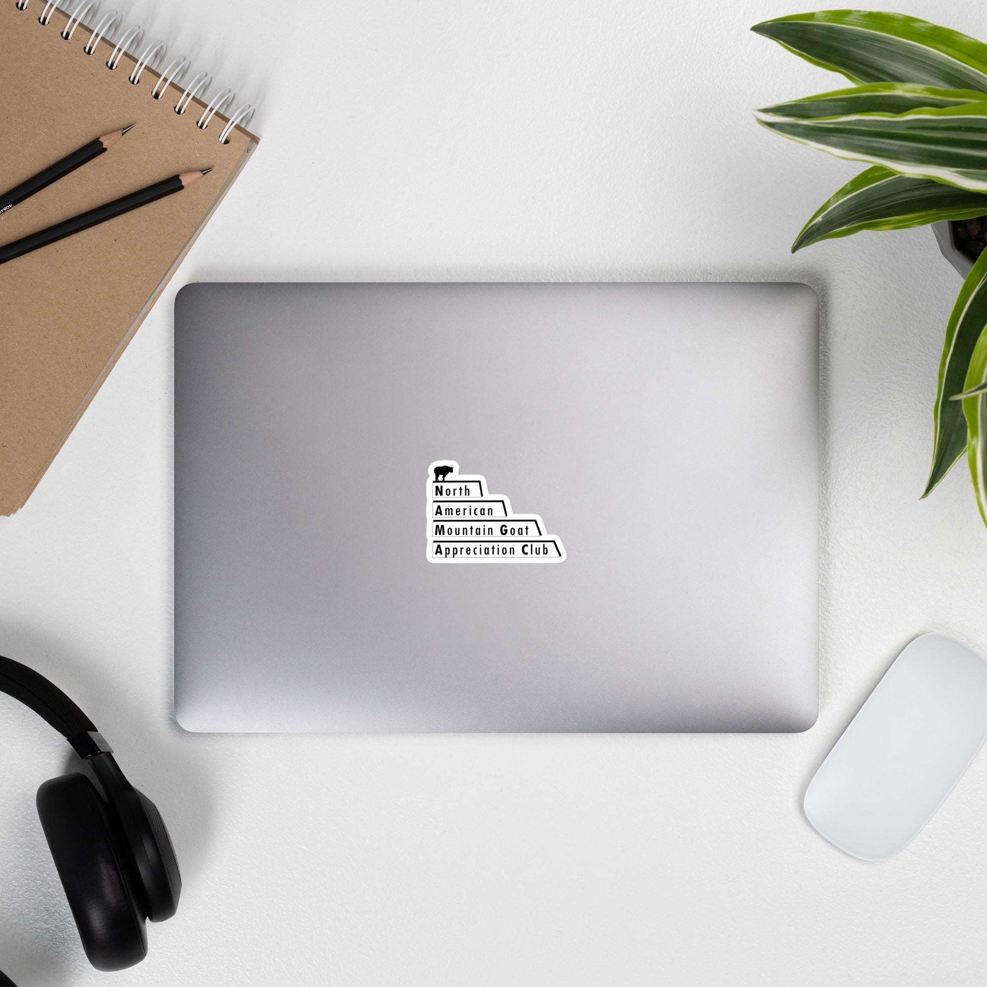 A laptop with a cover stick that is the North American Mountain Goat Appreciation Club logo, a pair of glasses, a pencil, a notebook, a mouse, and succulents