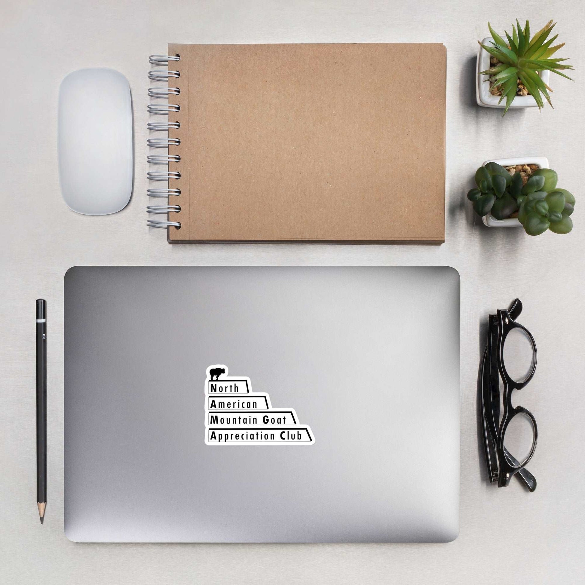 A laptop with a cover stick that is the North American Mountain Goat Appreciation Club logo, a pair of glasses, a pencil, a notebook, a mouse, and succulents