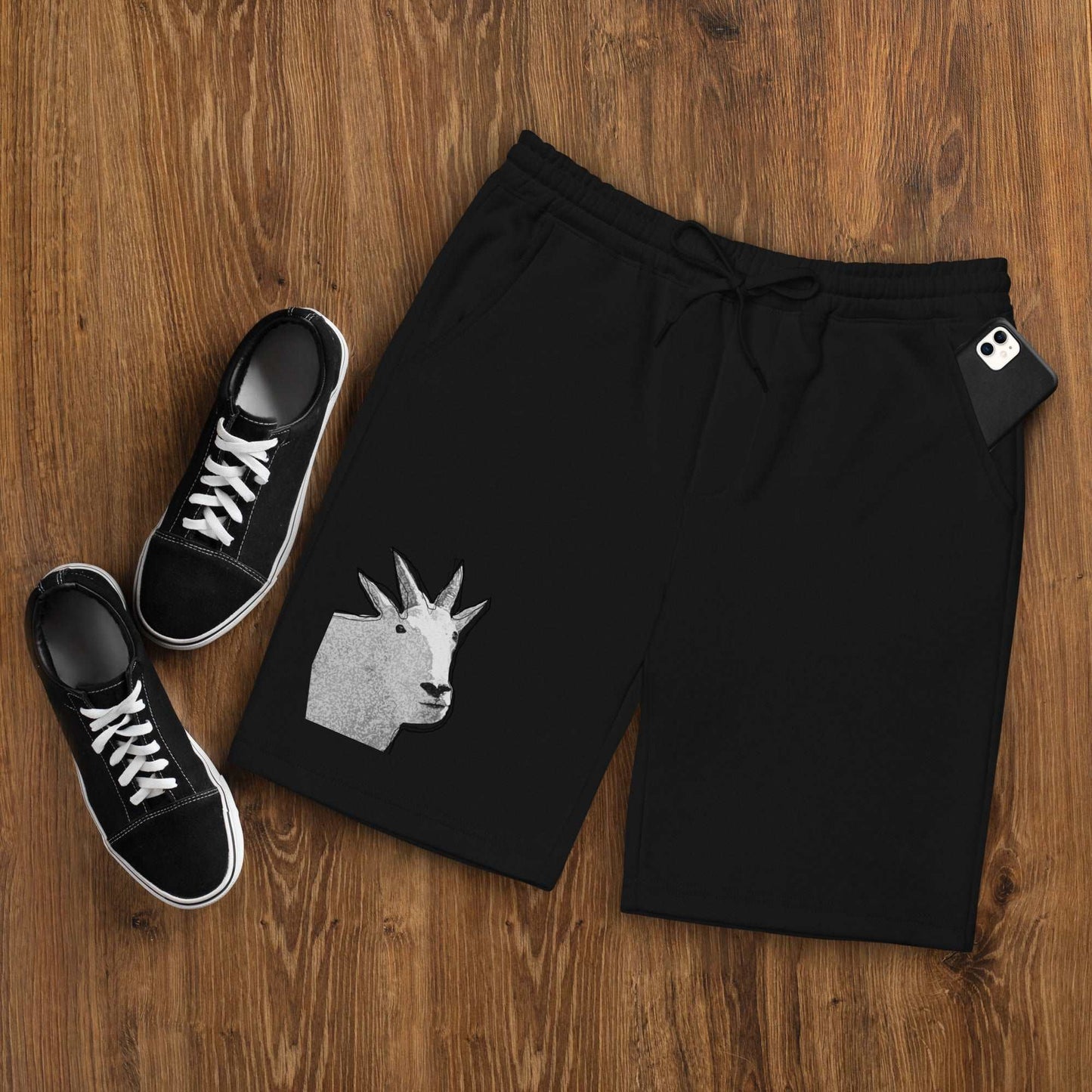 A pair of black Men's fleece shorts designed by North American Mountain Goat Appreciation Club that features a unique funny mountain goat design, placed on a natural wooden floor beside a pair of black sneakers.