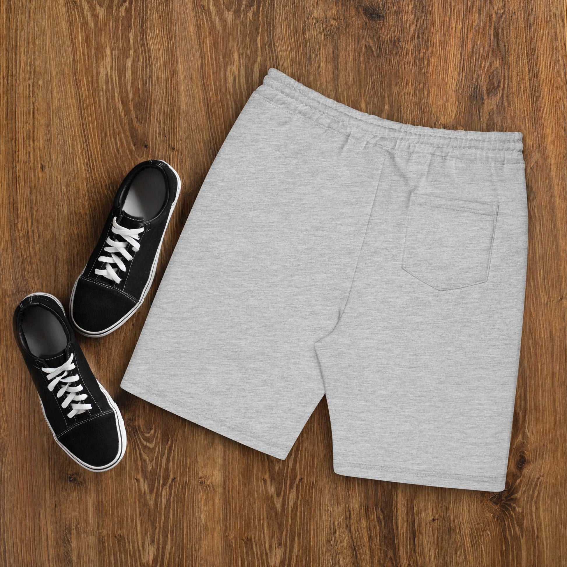 A pair of grey Men's fleece shorts designed by North American Mountain Goat Appreciation Club that features a unique funny mountain goat design, placed on a natural wooden floor beside a pair of black sneakers.