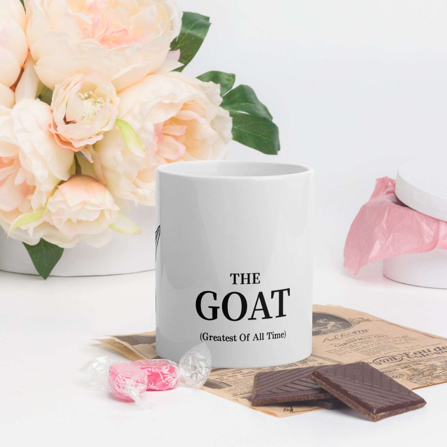 A white glossy mug that has a funny mountain goat design, the quote "The GOAT", and the logo North American Mountain Goat Appreciation Club.