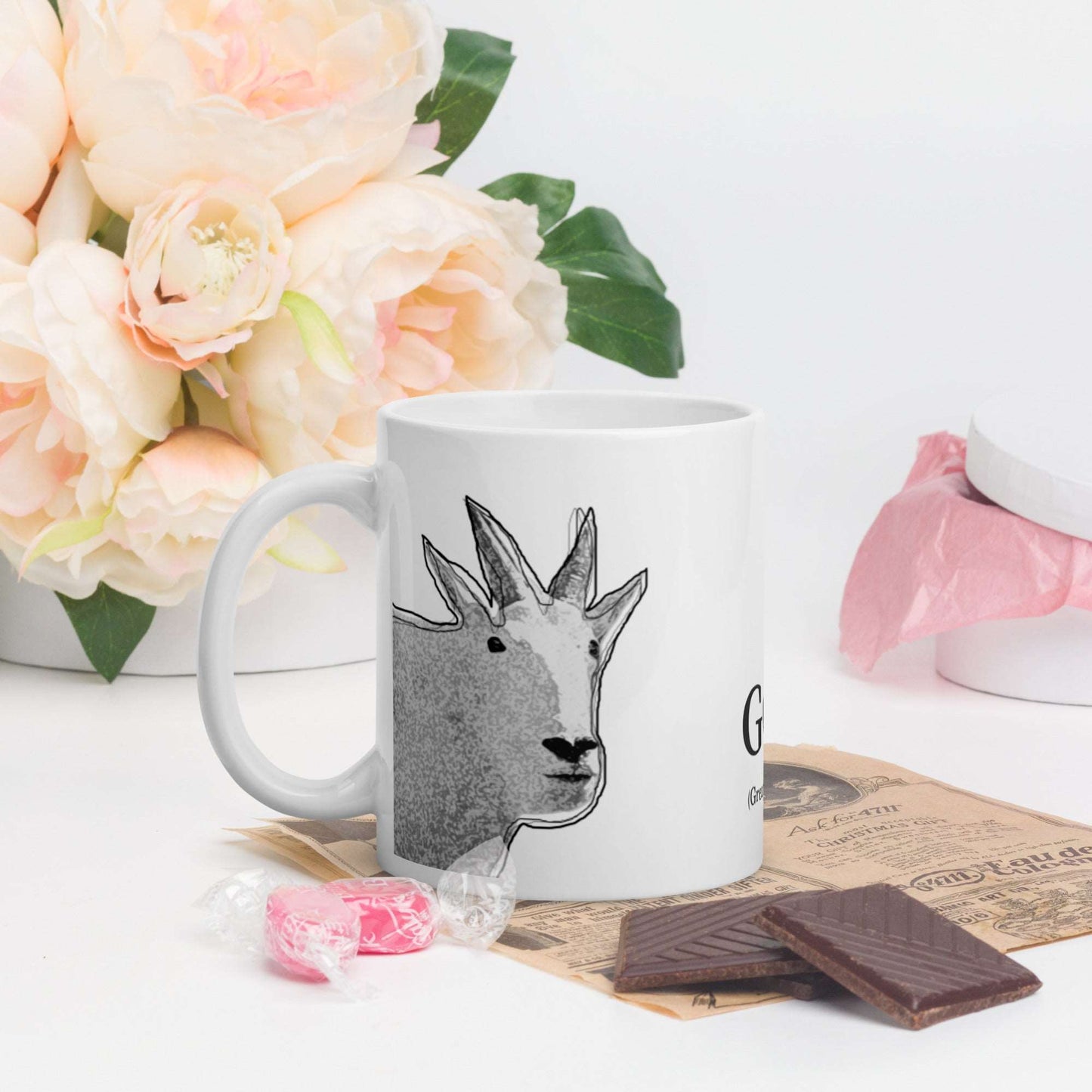 A white glossy mug that has a funny mountain goat design, the quote "The GOAT", and the logo North American Mountain Goat Appreciation Club.